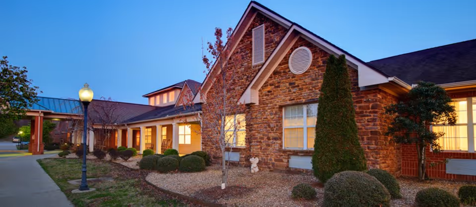 Stone-faced assisted living building front at dusk with lit windows, landscaped shrubs and a walkway.
