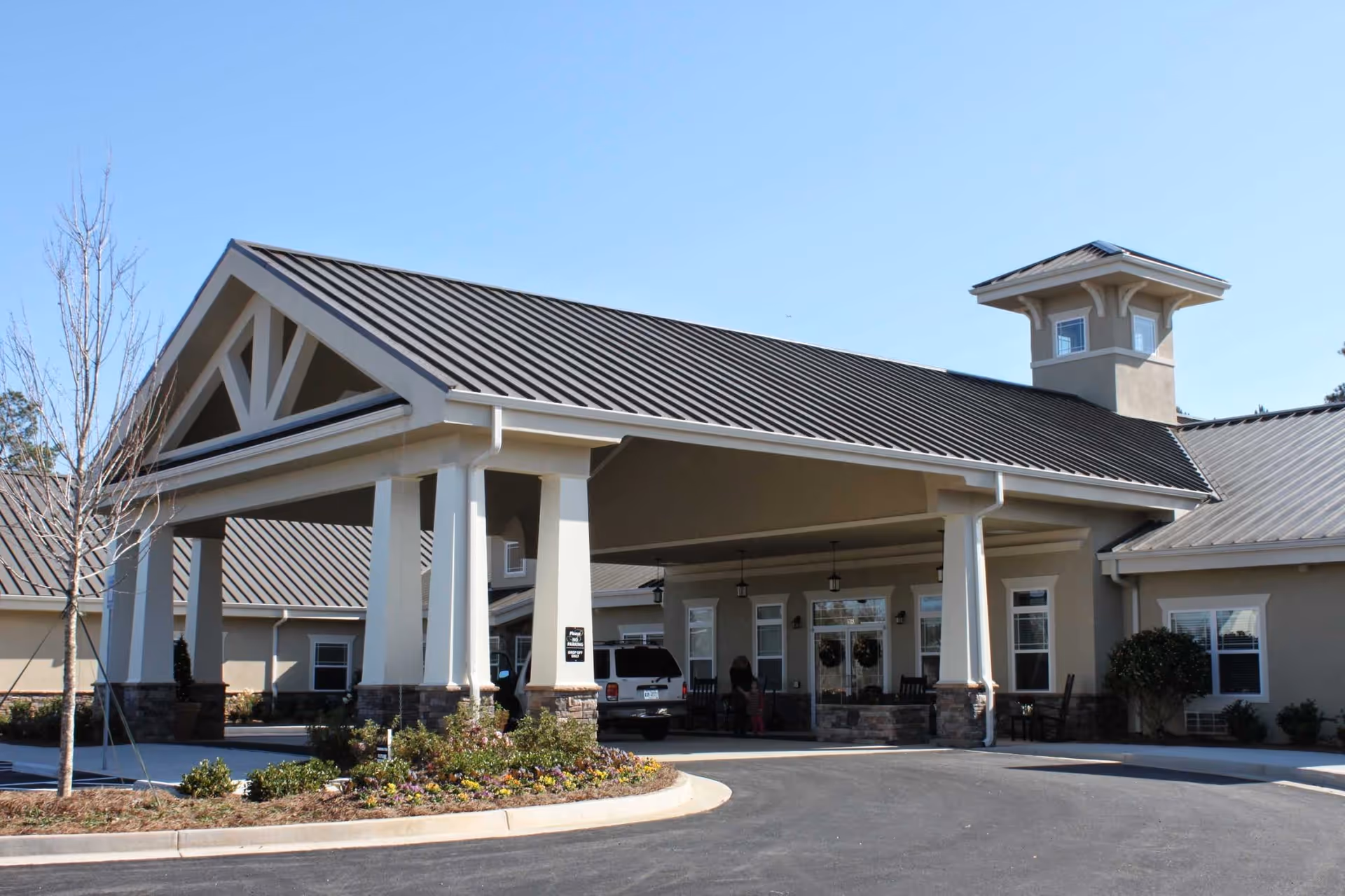 Exterior view of Benton House of Newnan showing the main entrance with a covered drop-off area supported by white columns, a vehicle parked under the canopy, and landscaping with flowers and shrubs in front.