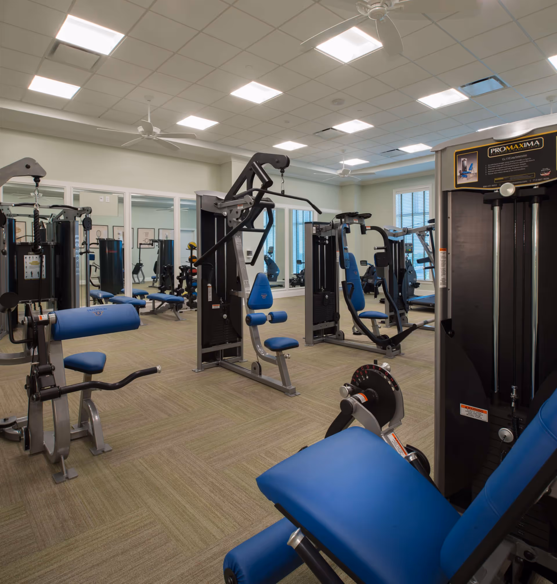 A well-lit fitness room with various exercise machines featuring blue padded seats and backrests. The room has a beige carpeted floor, white ceiling with recessed lighting, ceiling fans, and large windows with blinds. Mirrors line one wall, reflecting the equipment and creating a spacious feel.