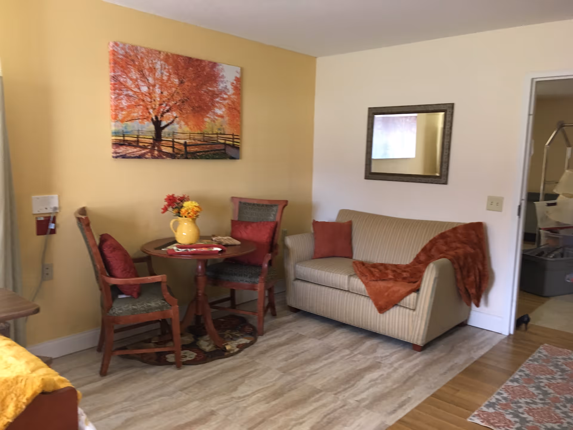 A cozy living area with a small round wooden table and two chairs with red cushions. A yellow vase with flowers is on the table. There is a beige striped loveseat with a red pillow and a red throw blanket. A framed mirror hangs on the wall above the loveseat, and a colorful painting of a tree with autumn leaves is on the adjacent wall. The floor has a mix of wood and tile, and an open doorway leads to another room.