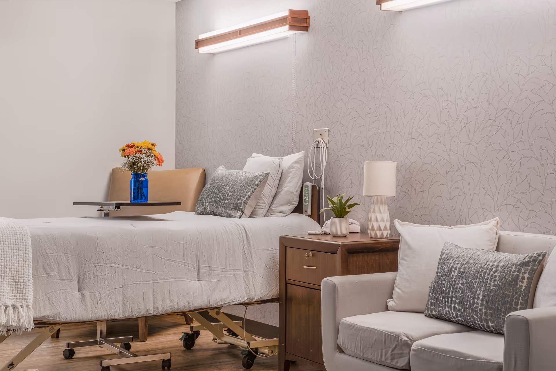A calm senior living bedroom with an adjustable bed, bedside table with lamp and flowers, and a cushioned armchair.