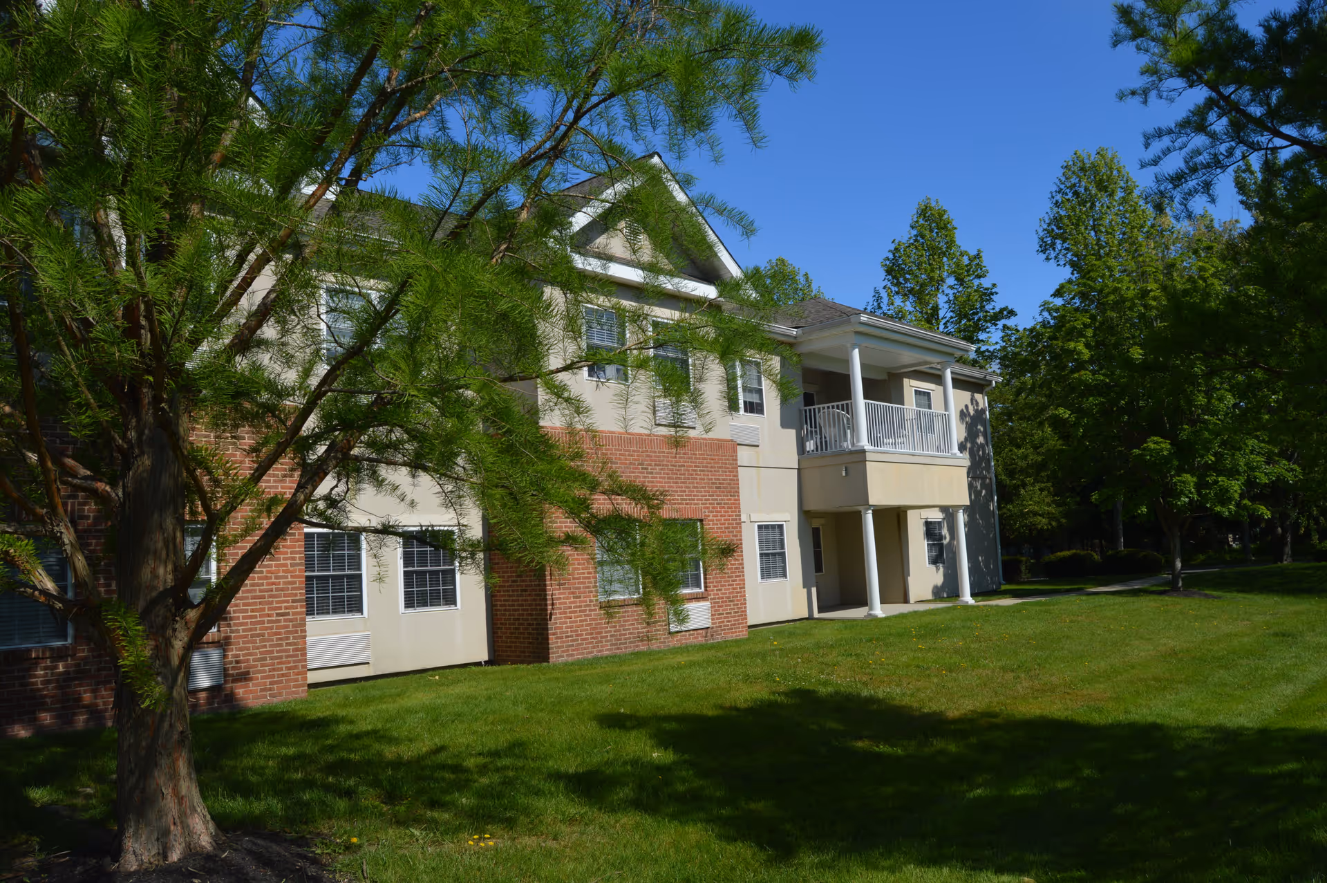 Exterior view of a two-story residential building with a combination of brick and beige siding, surrounded by green grass and trees under a clear blue sky.