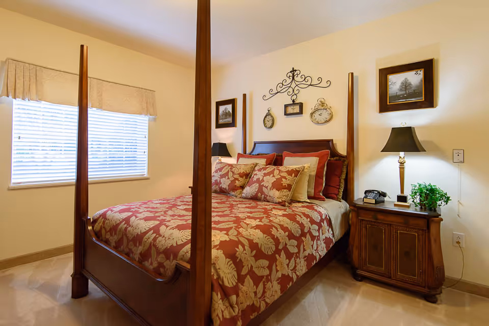 A bedroom with a wooden four-poster bed covered in a red and beige floral patterned bedspread and multiple pillows. There is a wooden nightstand with a lamp, a telephone, and a small plant. The walls are decorated with framed pictures and ornamental clocks. A window with blinds and a beige valance is on the left side.