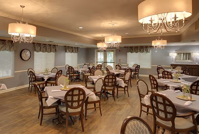A spacious dining room with multiple round tables covered with light-colored tablecloths, each set with napkins and small floral centerpieces. The room features wooden chairs with patterned upholstery, large windows with blinds and valances, elegant chandeliers hanging from the ceiling, and a wooden floor.