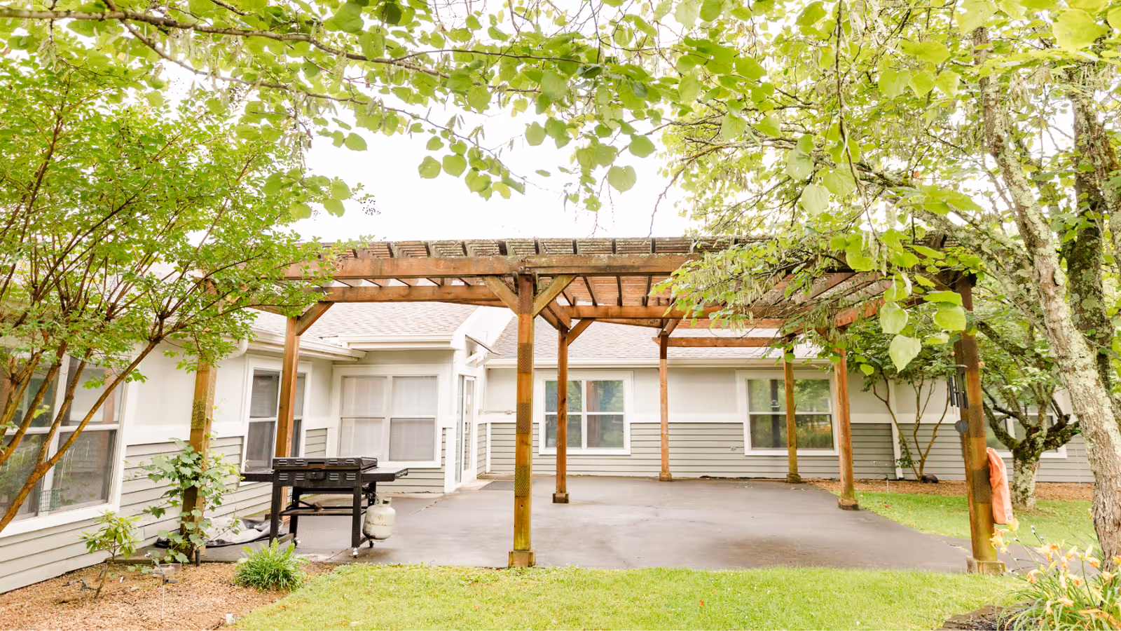 Outdoor patio area at Wexford House with a wooden pergola structure, surrounded by trees and greenery. The patio is paved and has a grill on one side. The building exterior is visible with windows and light-colored siding.