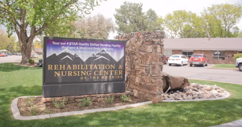 Outdoor view of a stone and metal sign for Rehabilitation & Nursing Center of the Rockies, situated on a grassy area with trees and a parking lot with cars in the background.
