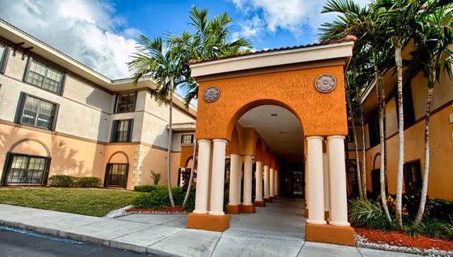 Exterior view of Presidential Place senior living facility showing a covered entrance with columns, palm trees, and a multi-story building with windows under a partly cloudy sky.