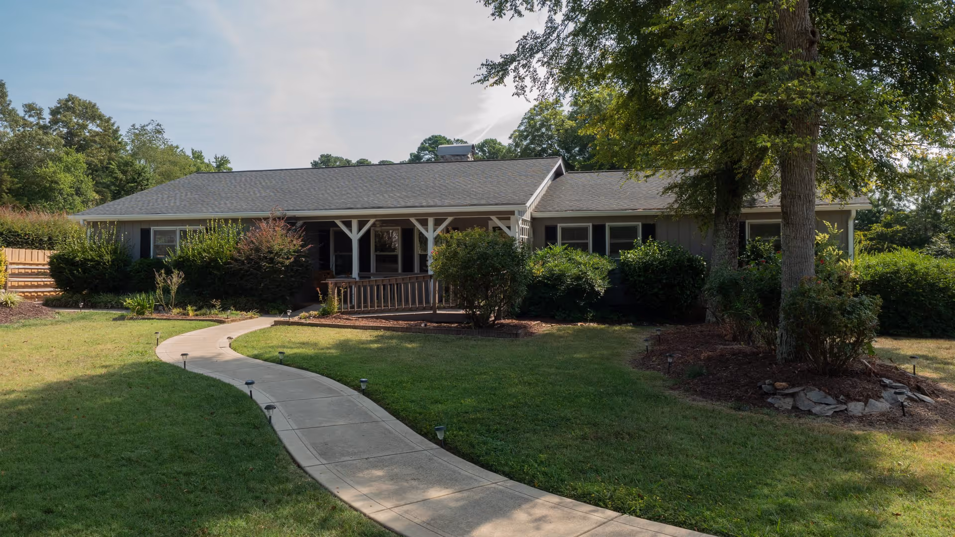 Single-story residential building with a gray roof and light-colored exterior surrounded by green bushes and trees. A curved concrete walkway leads from the foreground to the front porch of the building. The sky is clear with some light clouds.