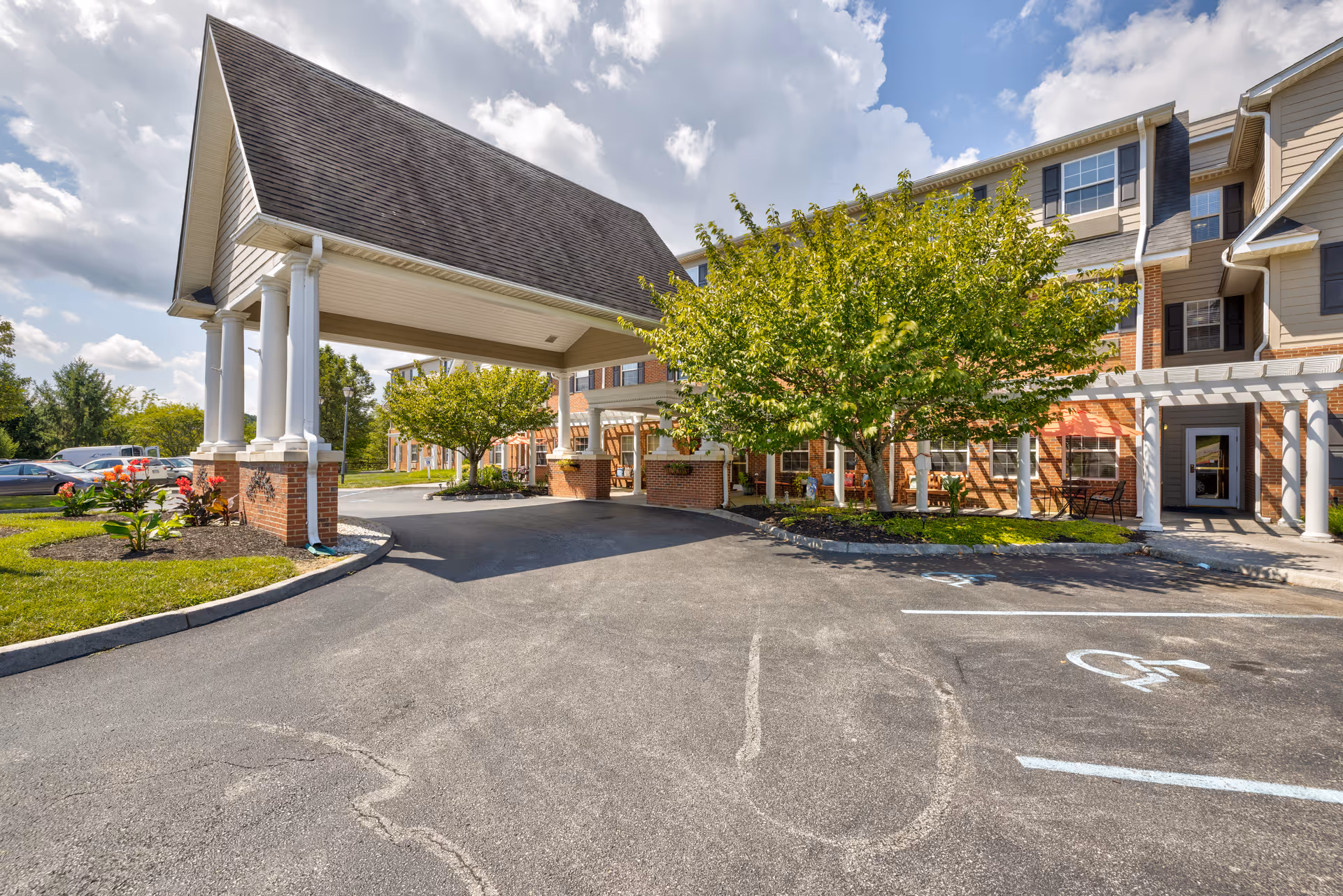 Entrance area of TerraBella Pheasant Ridge facility showing a covered driveway with white columns and a peaked roof. The building exterior features brick and siding with multiple windows. There are trees and landscaped areas with flowers near the entrance, and a parking lot with marked spaces including a handicapped parking spot.