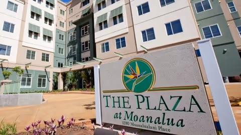 Exterior view of The Plaza at Moanalua building with a large sign in the foreground displaying the facility's name and address. The building is multi-story with multiple windows and a light-colored facade with green accents. There are plants and a paved area near the entrance.