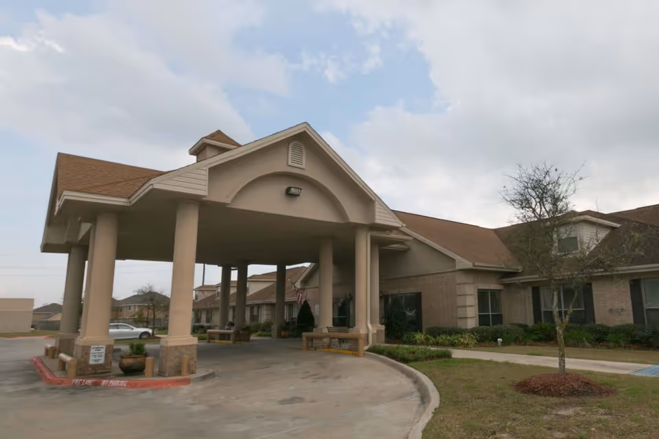 Exterior view of EastView HealthCare & Rehabilitation Center showing the main entrance with a covered driveway supported by large columns. The building has a beige facade with a brown roof, some landscaping with small trees and bushes, and a cloudy sky overhead.