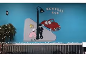 Interior wall painted teal with a mural depicting a girl in a pink dress and a boy in black silhouette standing near a red car under a street lamp. Above the mural, the words 'CENTRAL HUB' are displayed. Below the mural is a corrugated metal panel with string lights along the top edge, and a small decorated tree with white lights is visible on the left side.