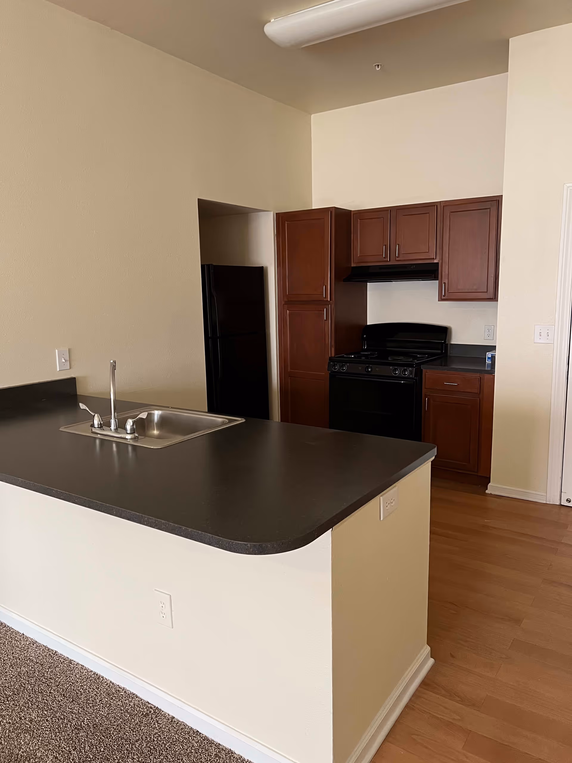 Interior view of a kitchen area featuring a black countertop with a built-in stainless steel sink and faucet. Behind the counter, there are wooden cabinets, a black stove with an overhead vent, and a black refrigerator partially visible in a recessed space. The floor is a combination of carpet and wood laminate, and the walls are painted a light beige color.