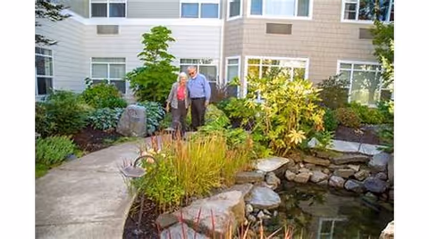 An elderly couple walking together on a curved concrete pathway through a landscaped garden area with various plants, shrubs, and a small pond with rocks, outside a multi-story building with large windows.