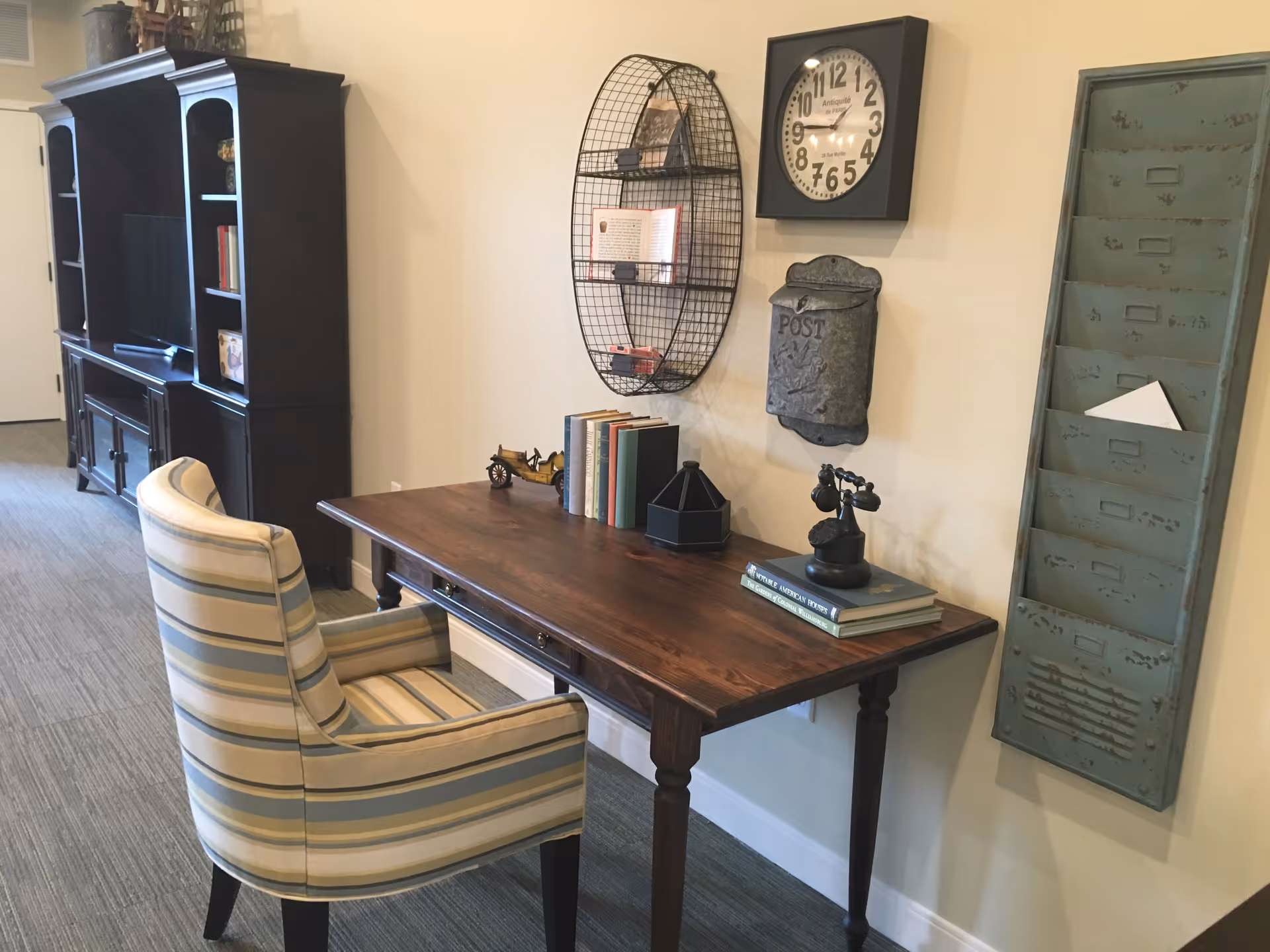 A wooden desk and striped upholstered chair in a decorated interior room with a wall clock, mail slots and shelving.