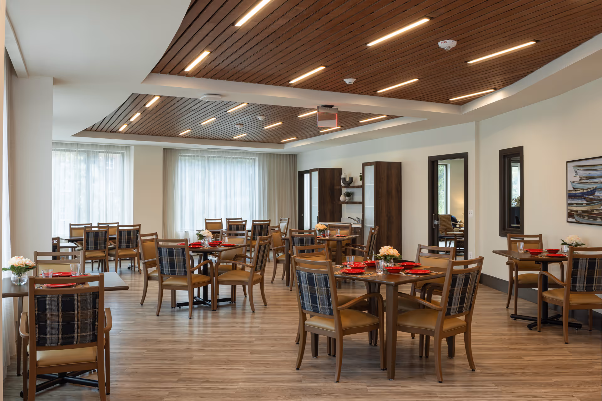 A spacious dining room with multiple wooden tables and chairs arranged neatly. Each table is set with red plates, glasses, and small flower arrangements. The room features large windows with sheer curtains allowing natural light to fill the space. The ceiling has wooden paneling with modern recessed lighting. There are cabinets and a painting on the walls, creating a warm and inviting atmosphere.