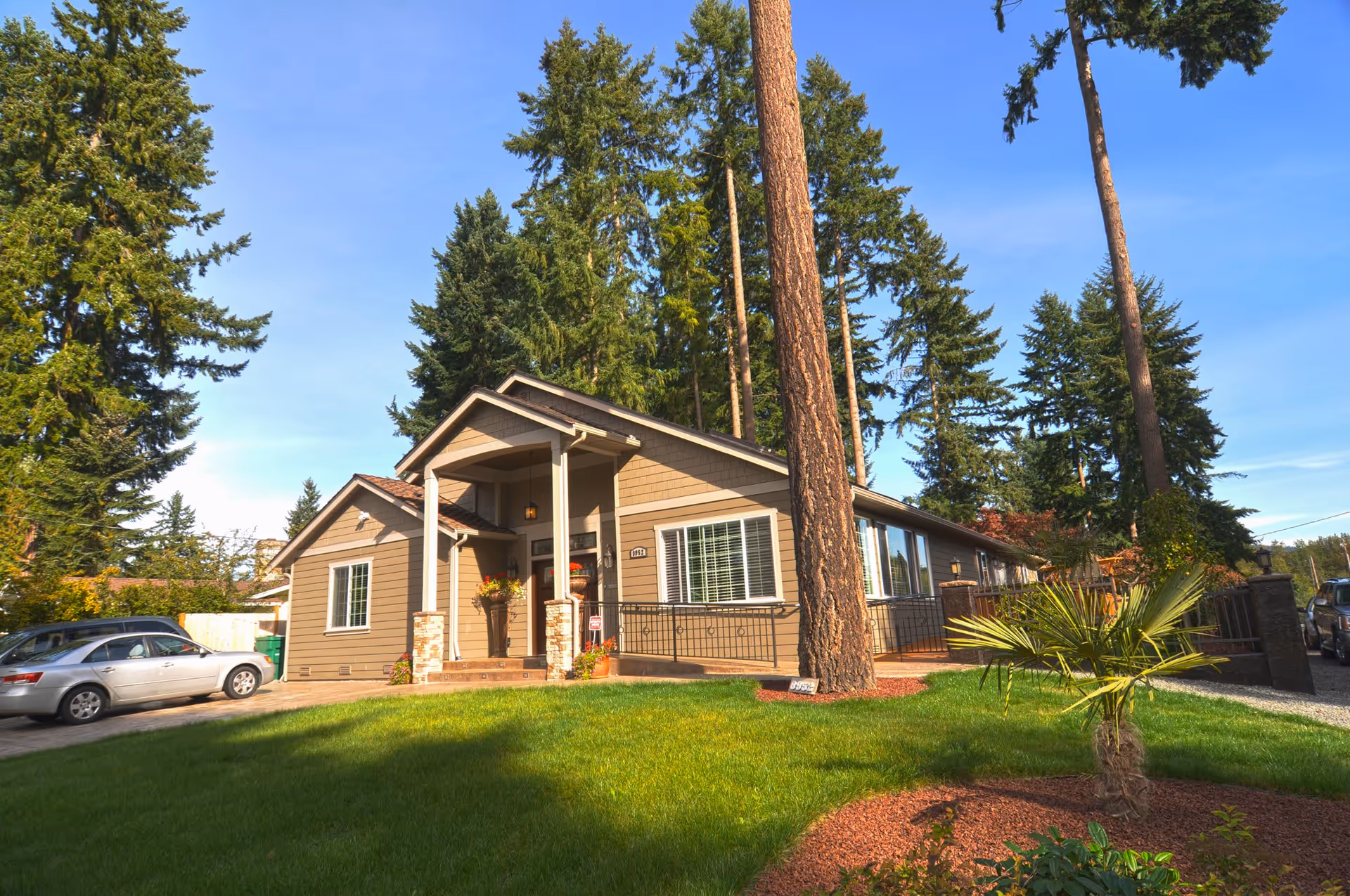 Exterior view of a single-story residential building with a covered front porch, surrounded by tall trees and a well-maintained lawn with landscaping. A silver car is parked on the driveway to the left of the house.