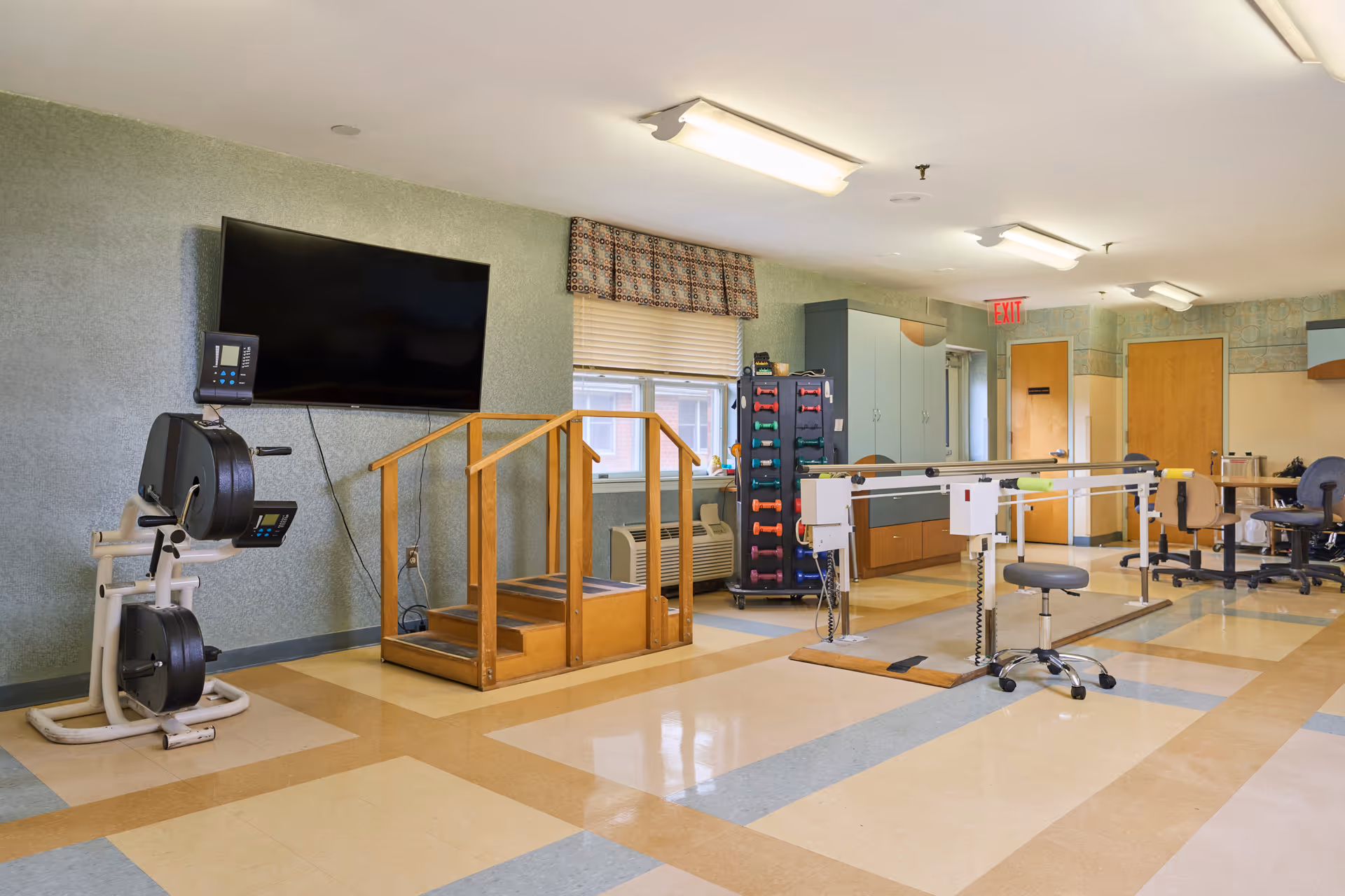 Rehabilitation/therapy room with exercise equipment, parallel bars, wooden therapy steps and a wall-mounted TV.