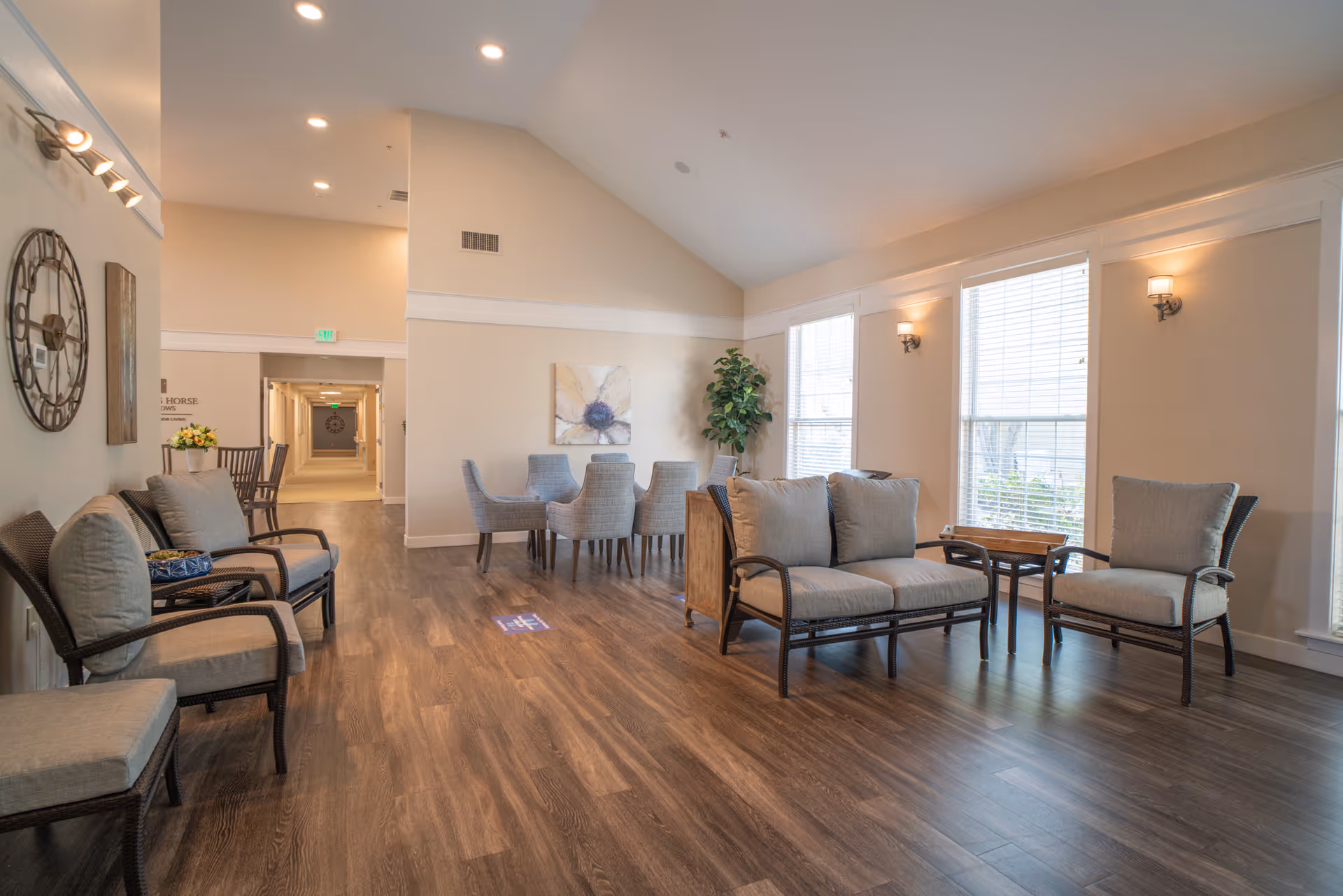 A spacious and well-lit common area in a senior living facility with comfortable seating including cushioned chairs and a loveseat arranged around small tables. The room features large windows with blinds, wooden flooring, wall sconces for lighting, a decorative wall clock, and a hallway leading to other parts of the facility. There is also a dining area with a table and chairs in the background.