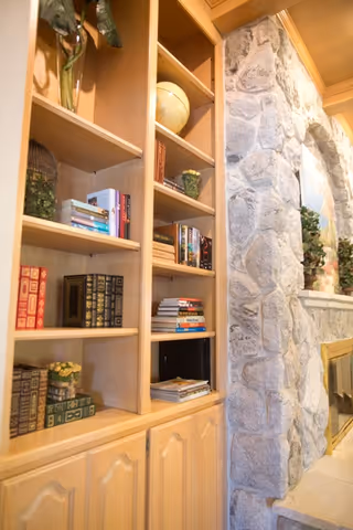 Wooden built-in bookshelf filled with books and decorative items next to a stone fireplace with a mantel adorned with plants and a framed picture above it.