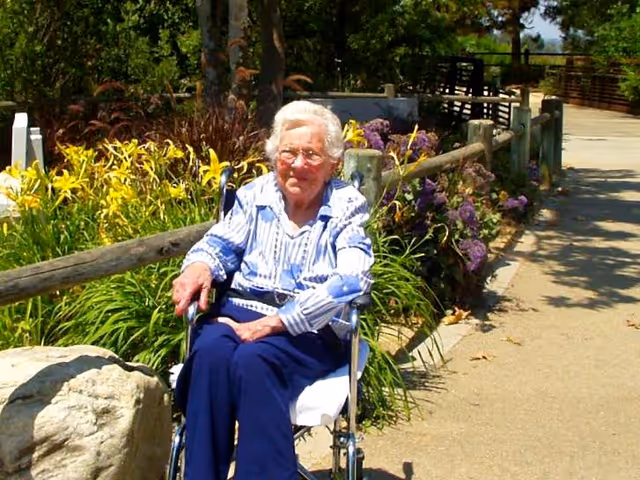 An elderly woman with white hair and glasses sitting in a wheelchair outdoors on a sunny day. She is wearing a blue and white striped shirt and dark blue pants. Behind her is a wooden fence with green plants and purple flowers along a paved pathway.