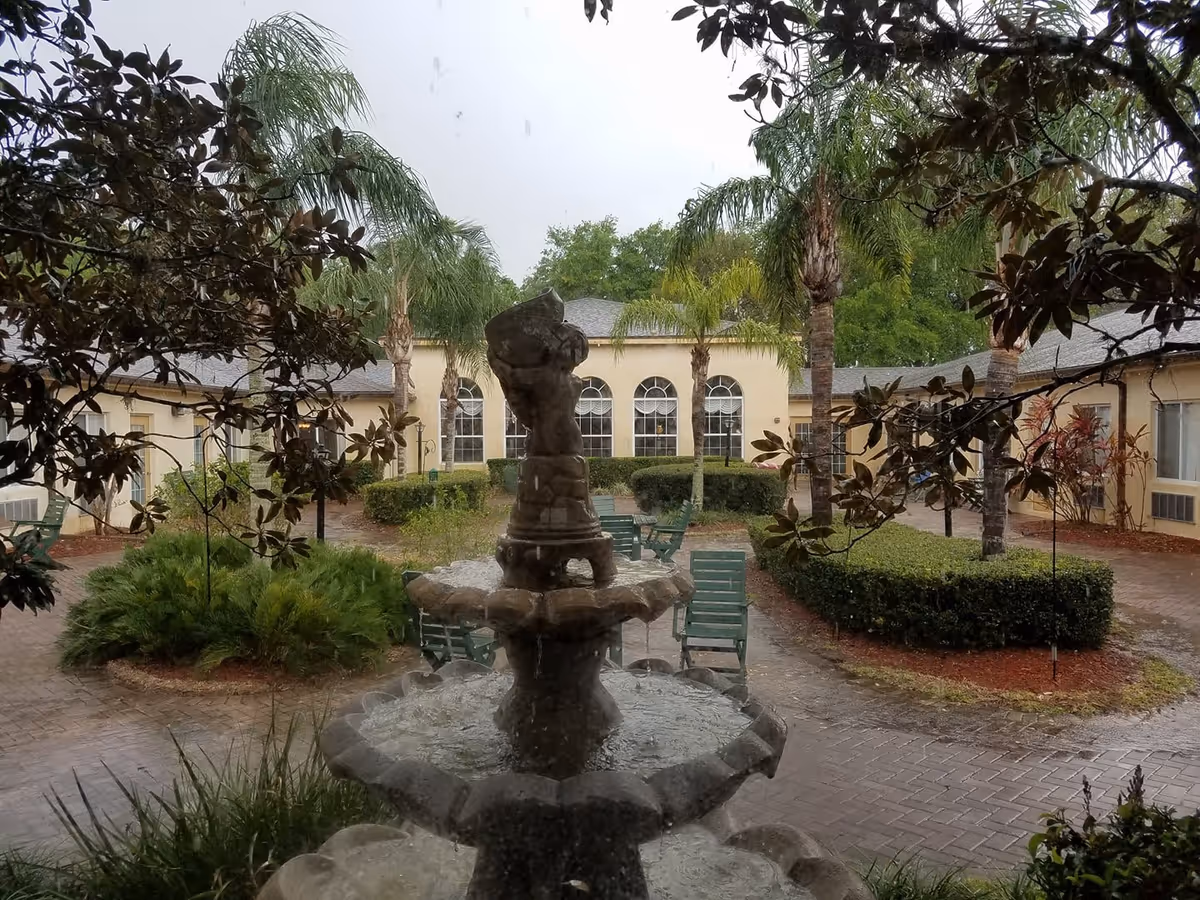 View of an outdoor courtyard at Savannah Court of Lakeland featuring a multi-tiered stone fountain in the center, surrounded by palm trees, green bushes, and several green chairs and tables. The courtyard is paved with bricks and enclosed by a beige building with arched windows.