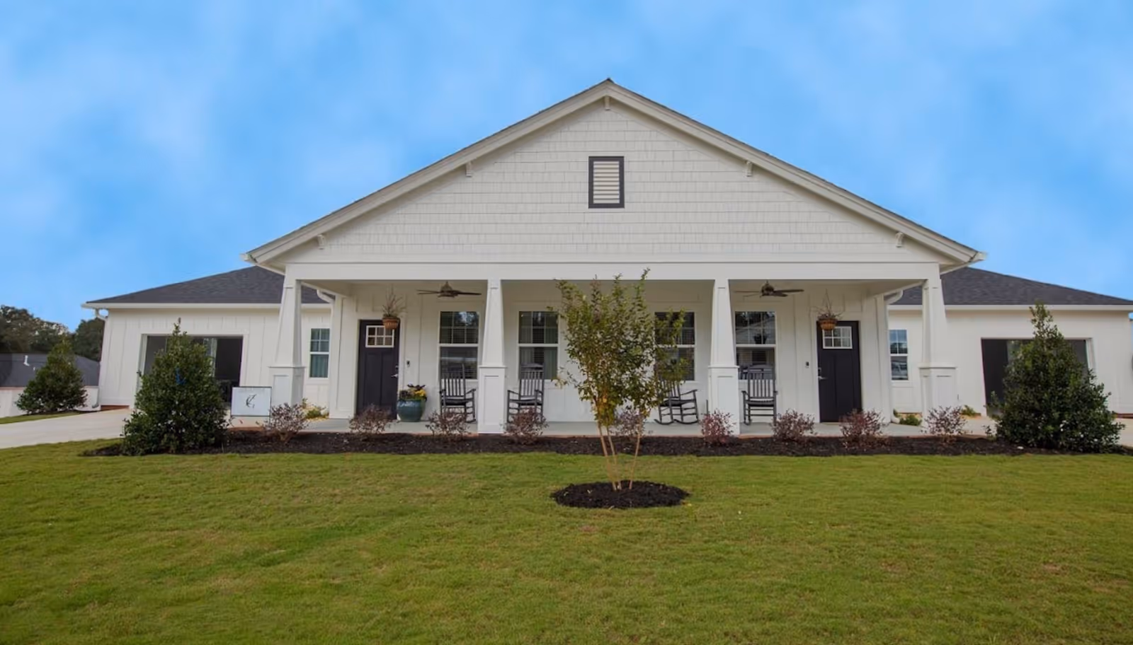 Front exterior view of a single-story building with a covered porch featuring rocking chairs, potted plants, and ceiling fans. The building is white with a gray roof, surrounded by a well-maintained lawn and small shrubs.