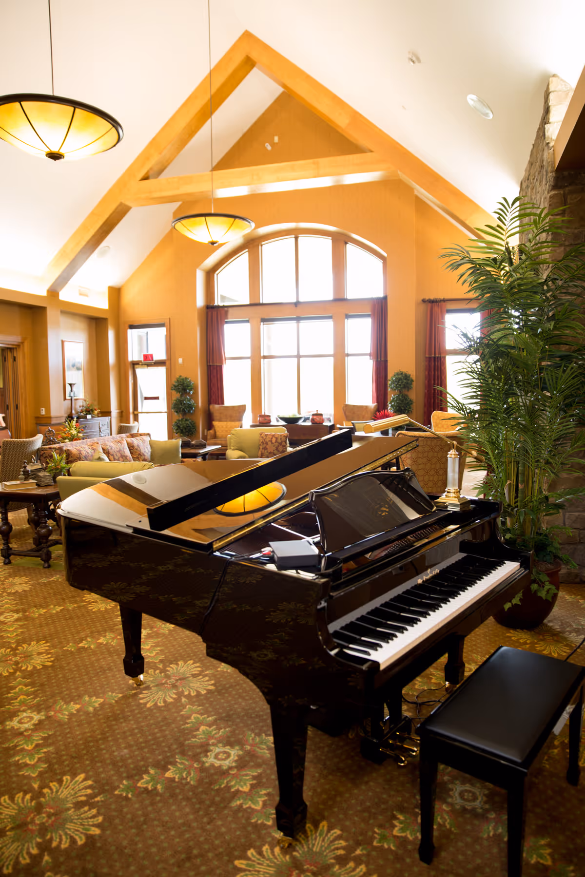 A spacious and warmly lit living room area with a grand piano in the foreground. The room features high vaulted ceilings with wooden beams, large windows with red curtains allowing natural light to fill the space, comfortable seating including sofas and armchairs, decorative plants, and elegant hanging light fixtures.