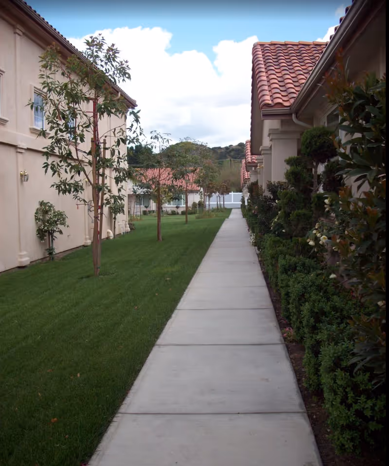 Concrete walkway between two residential buildings with red tile roofs, bordered by grass, trees, and shrubs.