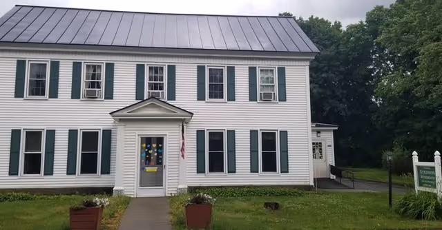 Front exterior view of a two-story white building with green shutters and a metal roof. The entrance has a small covered porch with an American flag hanging beside the door. There are two planters with flowers on either side of the walkway leading to the entrance. Trees and greenery surround the building.