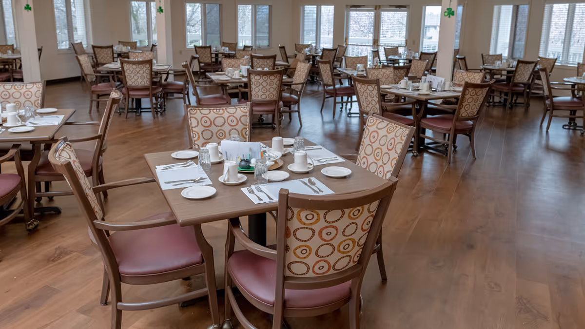 A spacious dining room in a senior living community with multiple wooden tables and chairs arranged neatly. Each table is set with white plates, cups, glasses, and silverware. The room has large windows allowing natural light to fill the space, and the floor is made of wood.