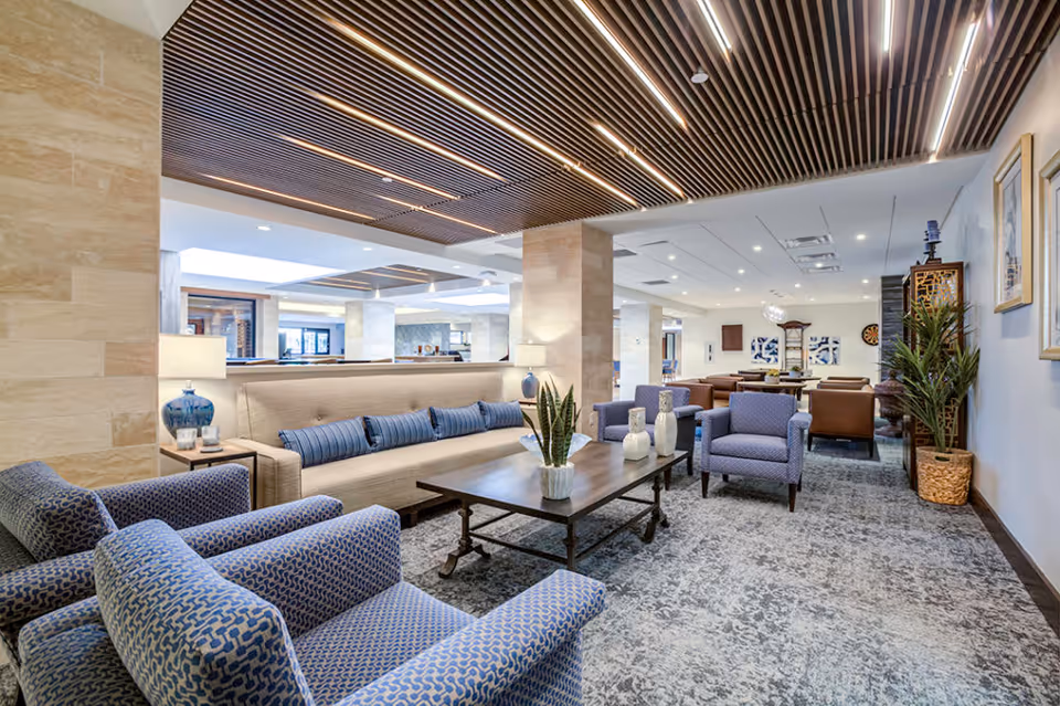 A spacious and well-lit living room area in The Palazzo facility featuring a beige sofa with blue cushions, two blue patterned armchairs, a wooden coffee table with decorative vases and a plant, and additional seating in the background. The ceiling has wooden slats with integrated lighting, and the floor is covered with a patterned carpet. There are plants and framed artwork on the walls, creating a comfortable and inviting atmosphere.