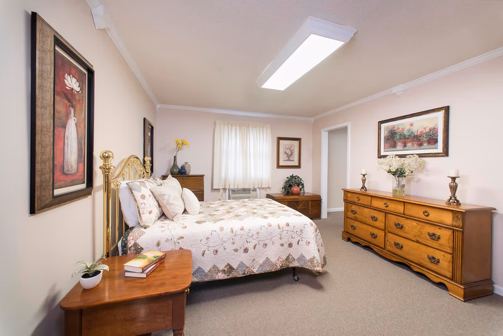 A cozy bedroom with a brass bed frame and a floral quilt. The room has beige walls and carpet, a wooden nightstand with books and a small plant, a wooden dresser with flowers and candles, and framed artwork on the walls. A window with sheer curtains lets in natural light.