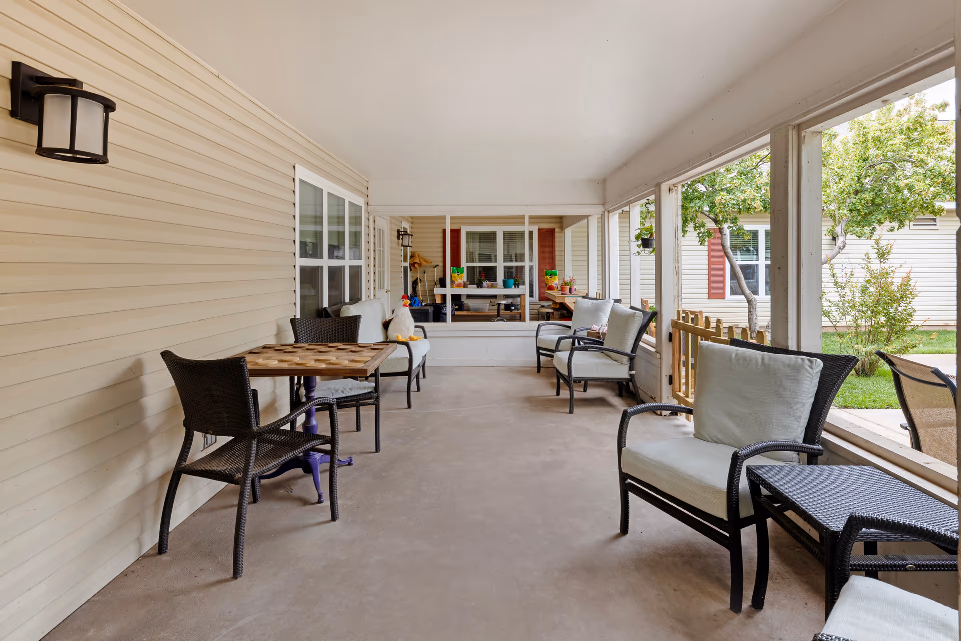 A covered outdoor patio area with several cushioned chairs and small tables. One table has a checkers board on it. The patio is adjacent to a beige building with windows and a door. There are plants and greenery visible outside the patio area.