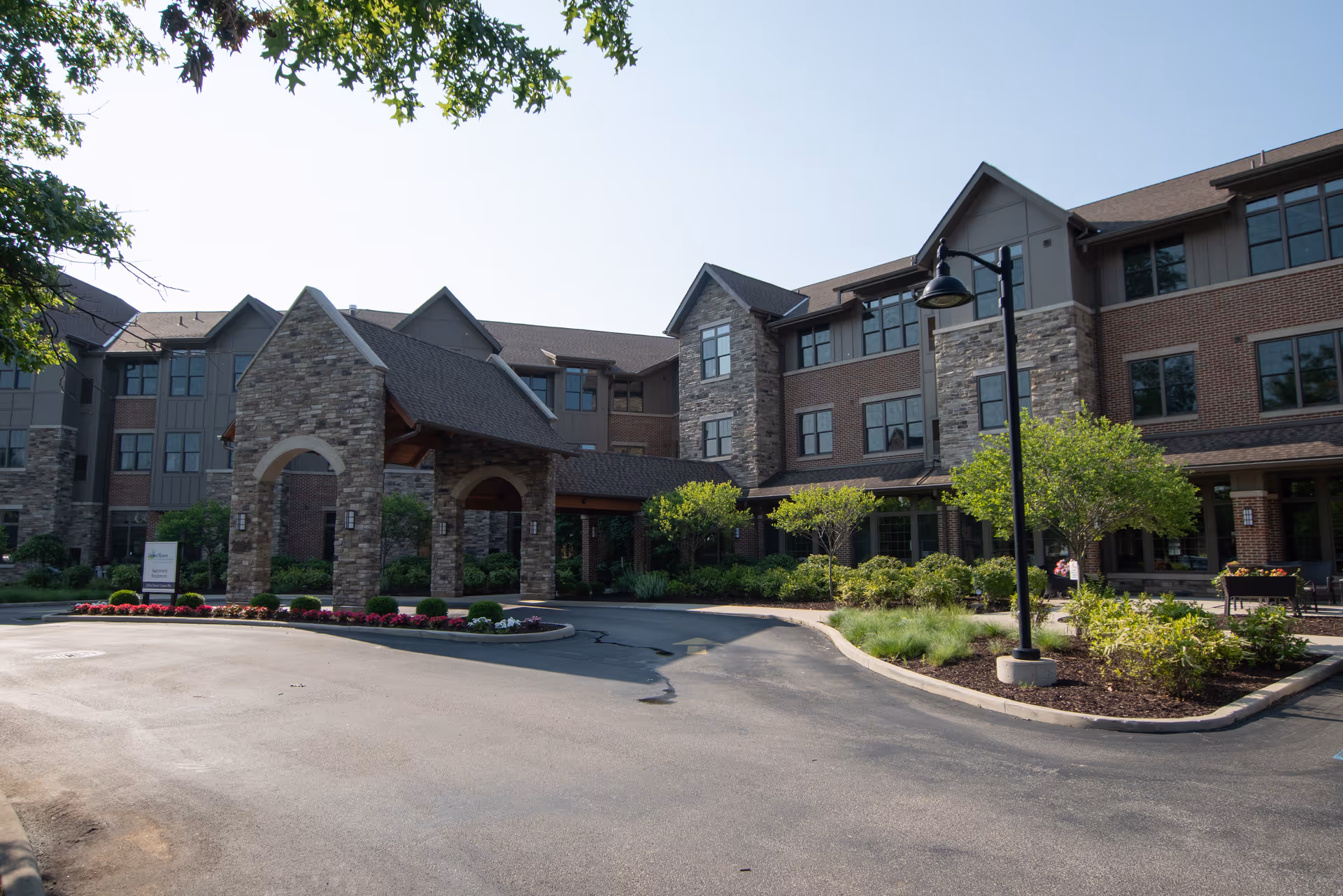 Front entrance of a three-story brick and stone senior living building with a covered porte-cochere and landscaped driveway.