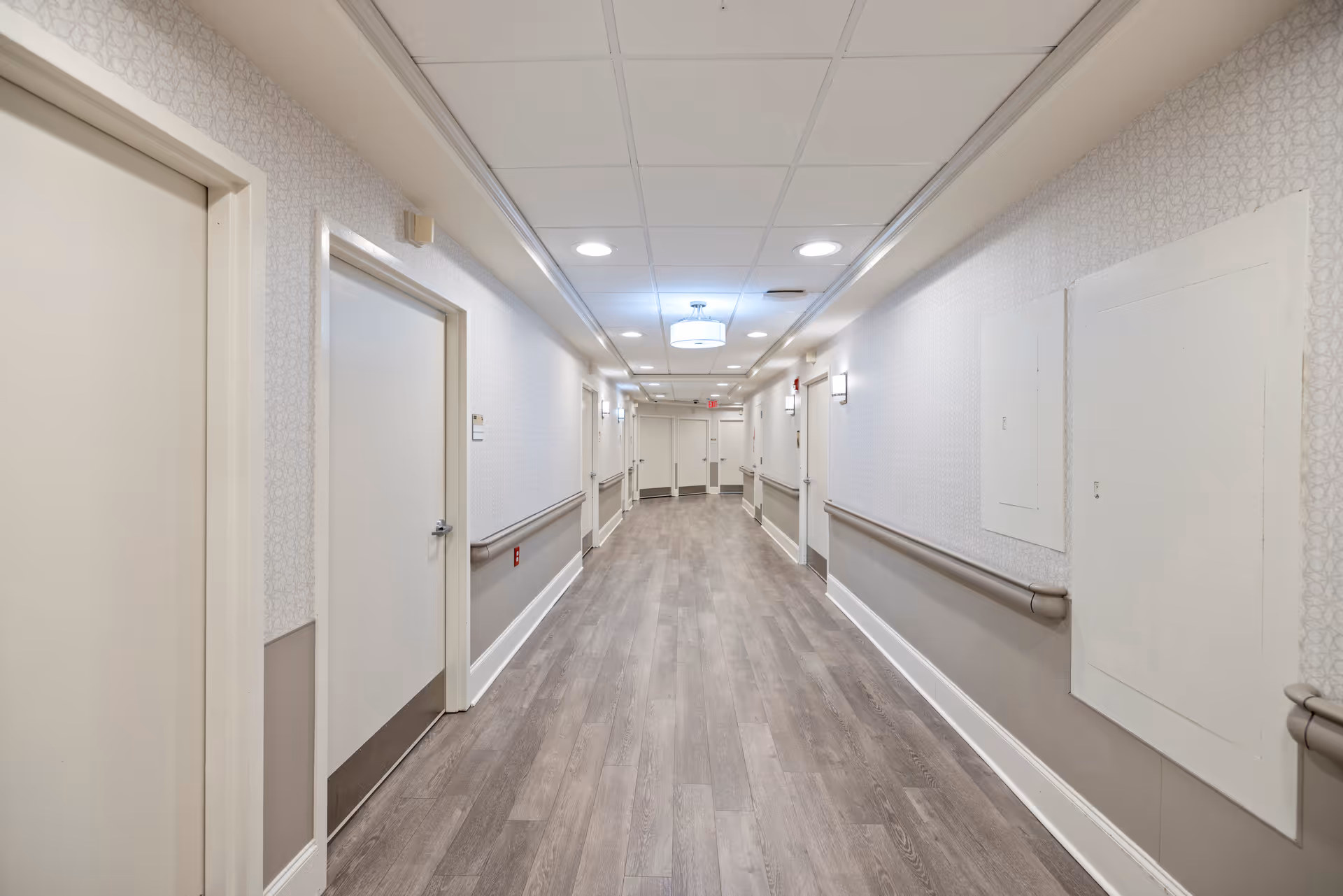 A long, well-lit hallway in a senior living facility with light-colored walls, wood flooring, multiple closed doors on both sides, handrails along the walls, and ceiling lights.