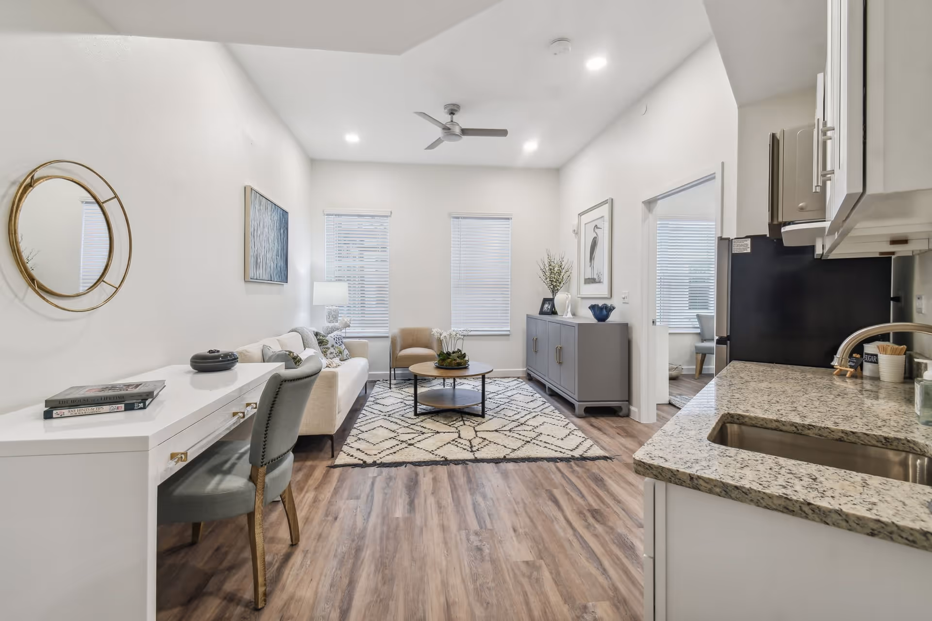 A bright and modern living area with light wood flooring, a white sofa, a beige armchair, a round coffee table on a patterned rug, a gray sideboard with decorative items, and a ceiling fan. To the right is a kitchen area with granite countertops and a stainless steel sink. A white desk with a gray chair and books is on the left side of the room. Two windows with blinds allow natural light into the space.