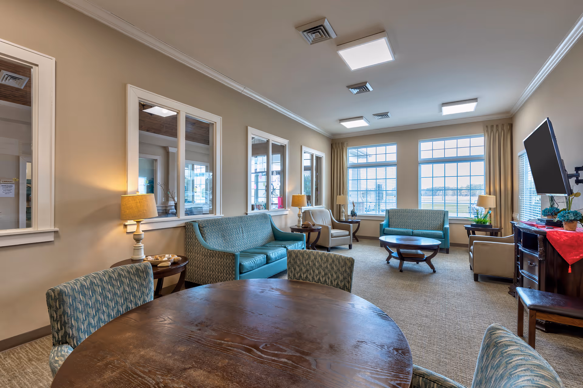A bright and spacious living room area with large windows letting in natural light. The room features two teal sofas, two beige armchairs, a round wooden coffee table, and a round wooden dining table with patterned chairs. There are multiple table lamps, a flat-screen TV mounted on the wall, and decorative plants on side tables. The walls are painted beige with white trim, and the carpet is light brown.