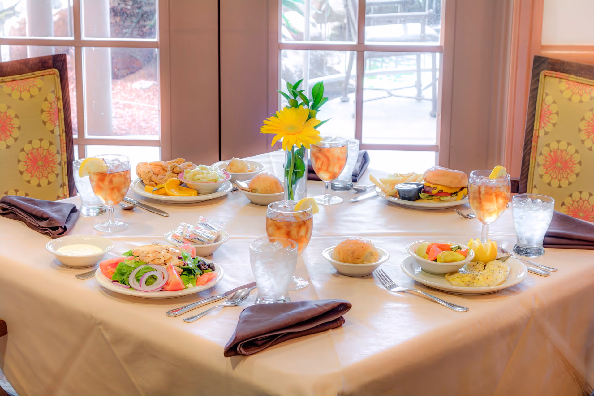 A dining table set for four with plates of food including salad, fried chicken with sides, a cheeseburger with fries, and a fish fillet with vegetables. Each place setting has a glass of iced tea with lemon, a glass of water, and a folded dark napkin. A small vase with a yellow flower is in the center of the table. The background shows a window with a view of an outdoor patio area.