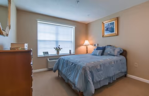 A cozy bedroom with a neatly made bed covered in a blue bedspread and multiple pillows. There is a wooden dresser with a mirror on the left side, a window with blinds letting in natural light, a small air conditioning unit below the window, a table lamp on a nightstand beside the bed, and a framed picture hanging on the wall above the bed.
