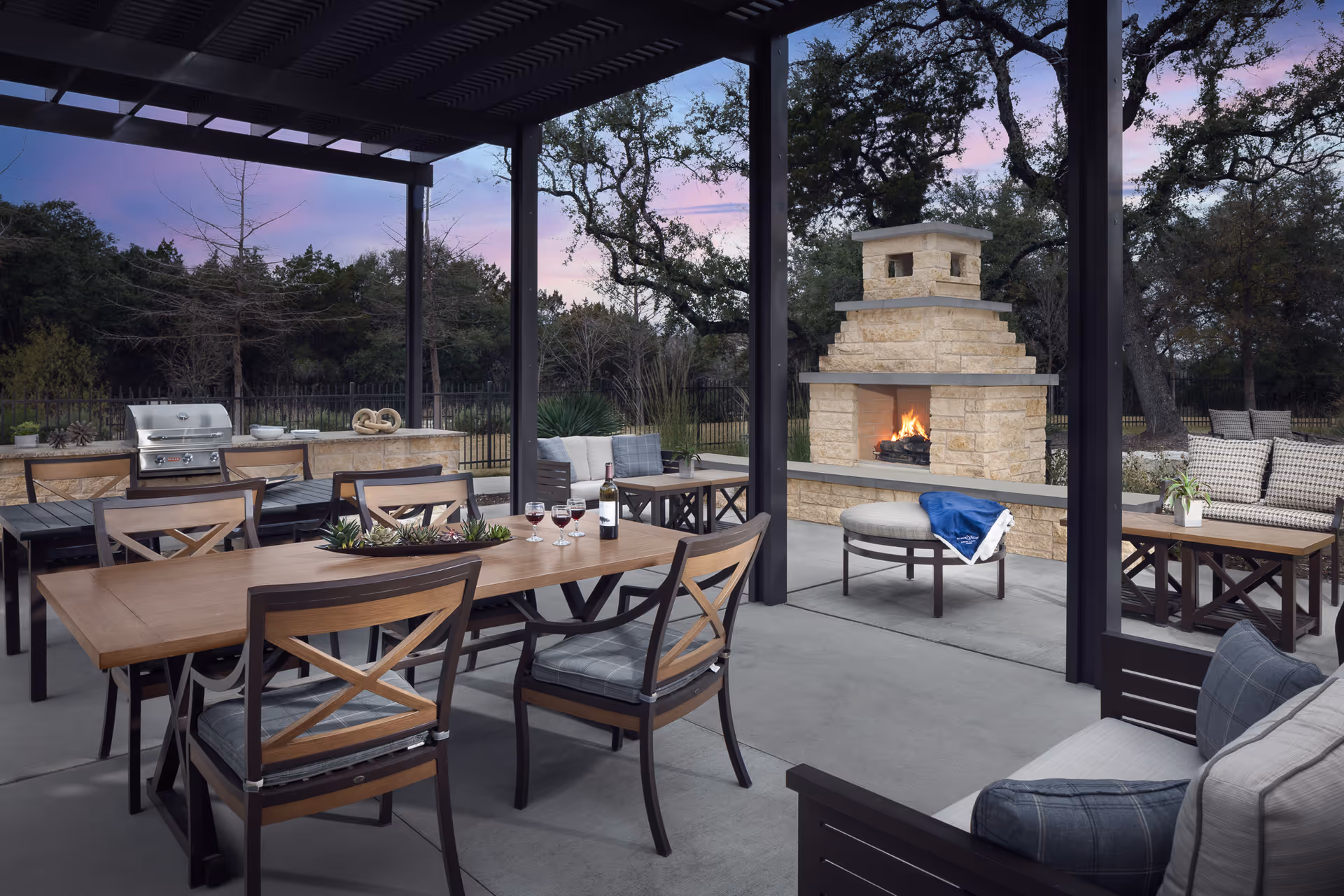 Outdoor covered patio area with wooden tables and chairs, a stone fireplace with a fire burning, cushioned seating, a grill, and trees in the background during dusk.