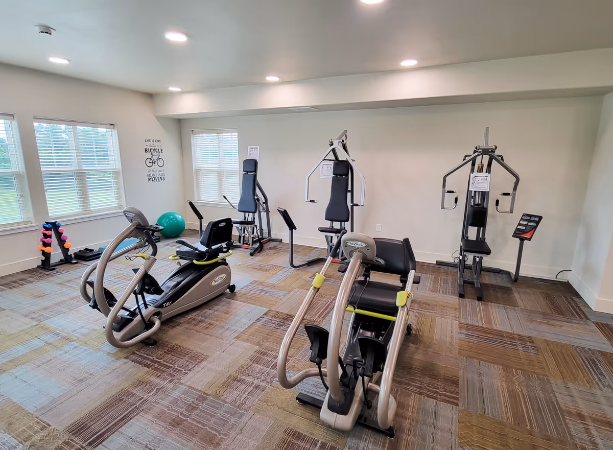 A well-lit exercise room with various fitness equipment including two NuStep machines, two weight machines, a rack of colorful dumbbells, and a green exercise ball. The room has large windows with blinds, beige walls, and a patterned carpet floor. A motivational wall decal reads 'Life is like riding a bicycle. To keep your balance you must keep moving.'