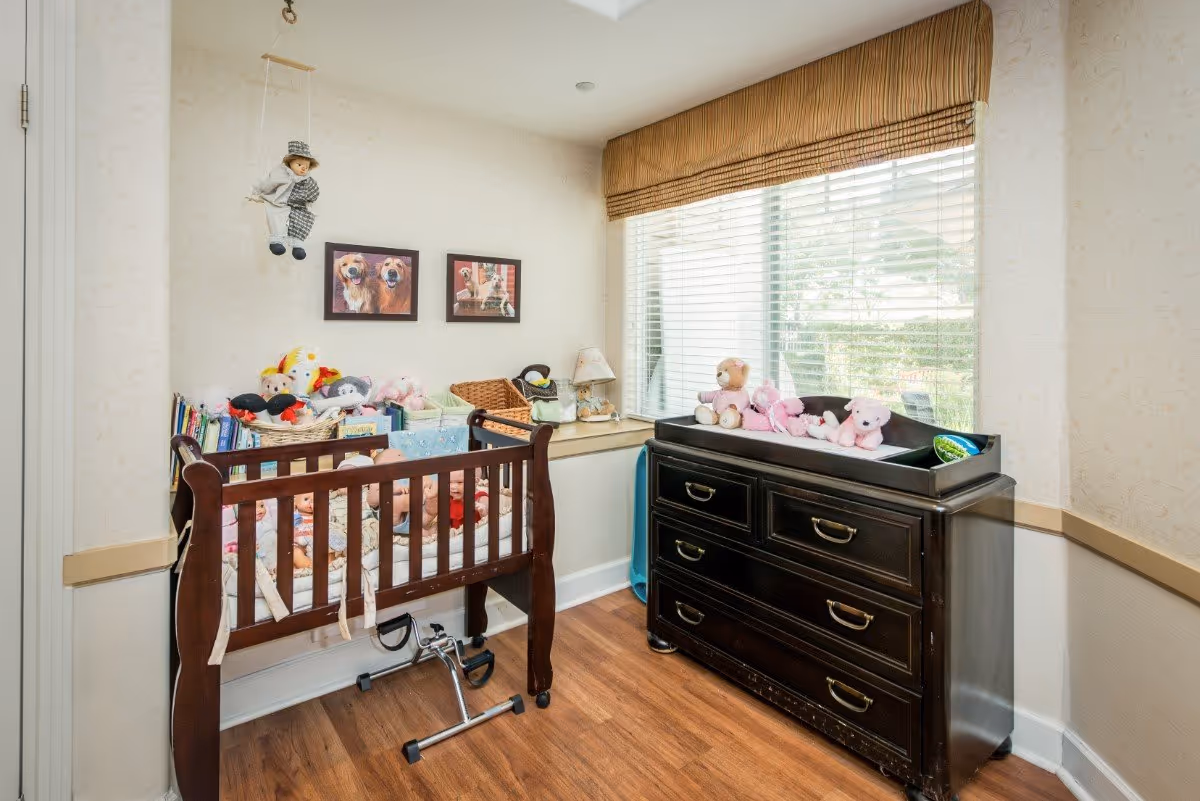 Nursery-style bedroom with a wooden crib filled with stuffed animals and a dark dresser changing table beneath a large window.