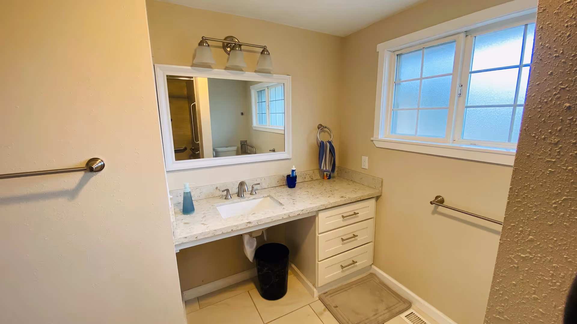 Bathroom with a marble-topped vanity, sink and mirror, towel racks, frosted window and overhead light fixture.