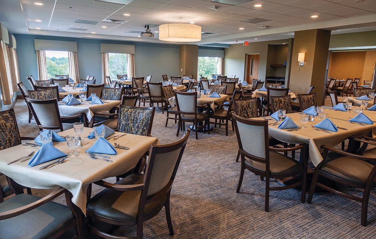 A spacious dining room with multiple tables covered in beige tablecloths, each set with blue folded napkins, glassware, and silverware. The room has large windows with views of greenery outside, patterned carpet flooring, and a modern ceiling light fixture.