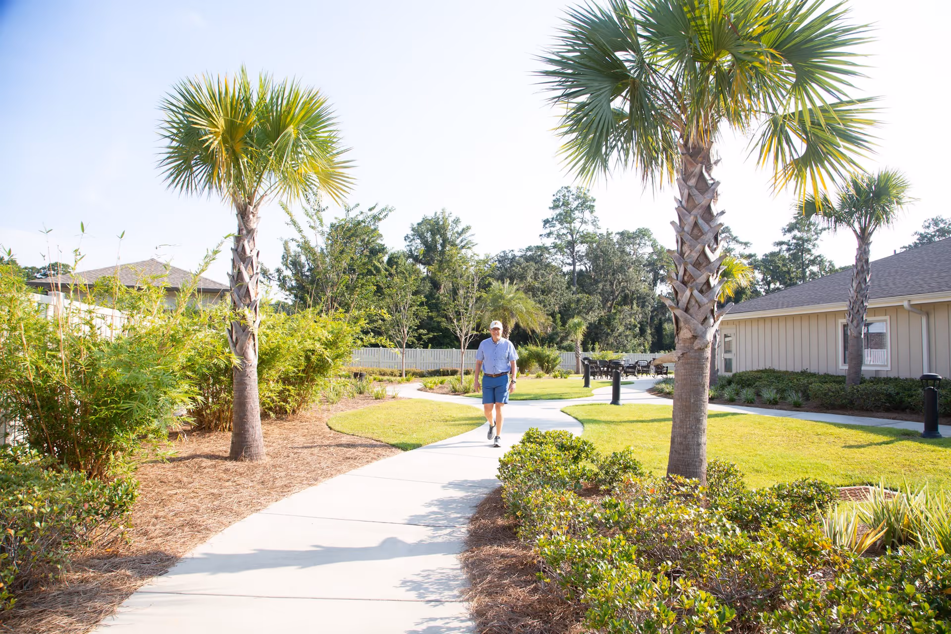 A man walking on a curved concrete pathway in a landscaped outdoor area with palm trees, bushes, and grass. There are buildings on the right side and a white fence in the background under a clear blue sky.
