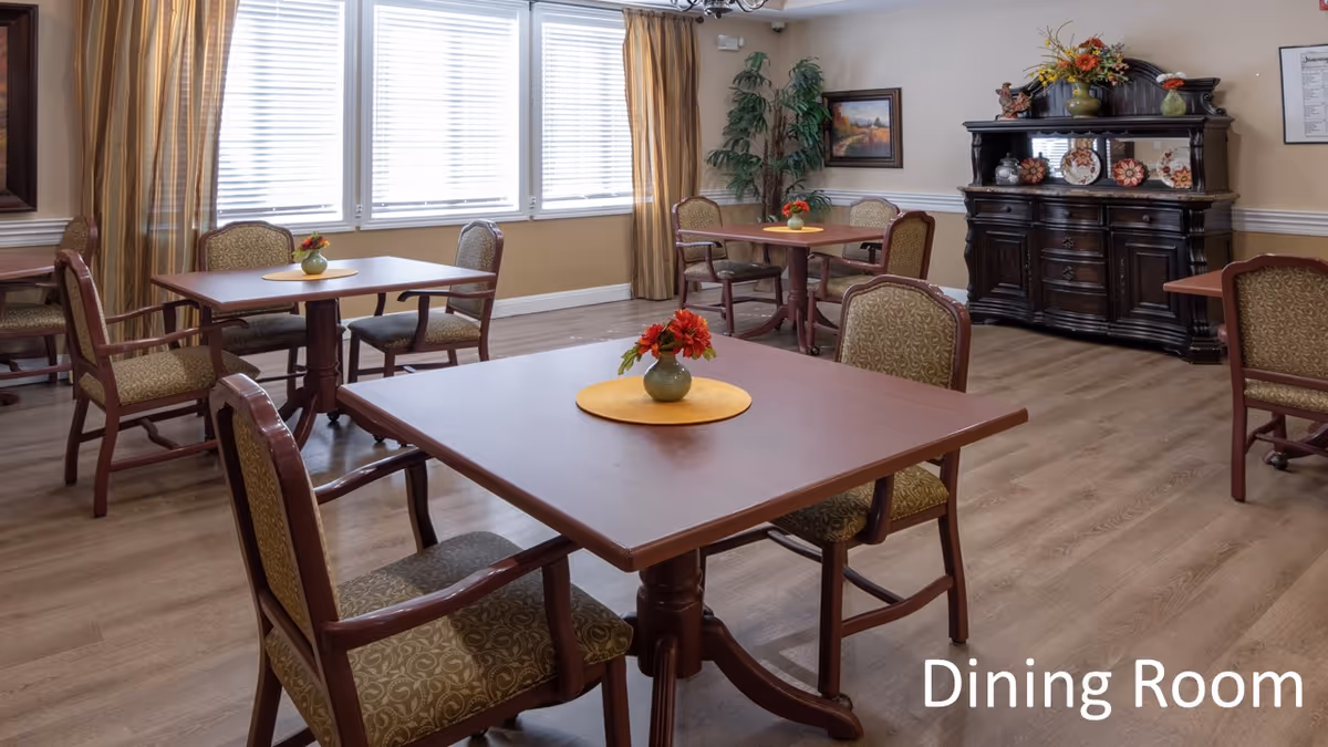Bright dining room with multiple wooden tables and upholstered chairs, small floral centerpieces, and a dark sideboard.