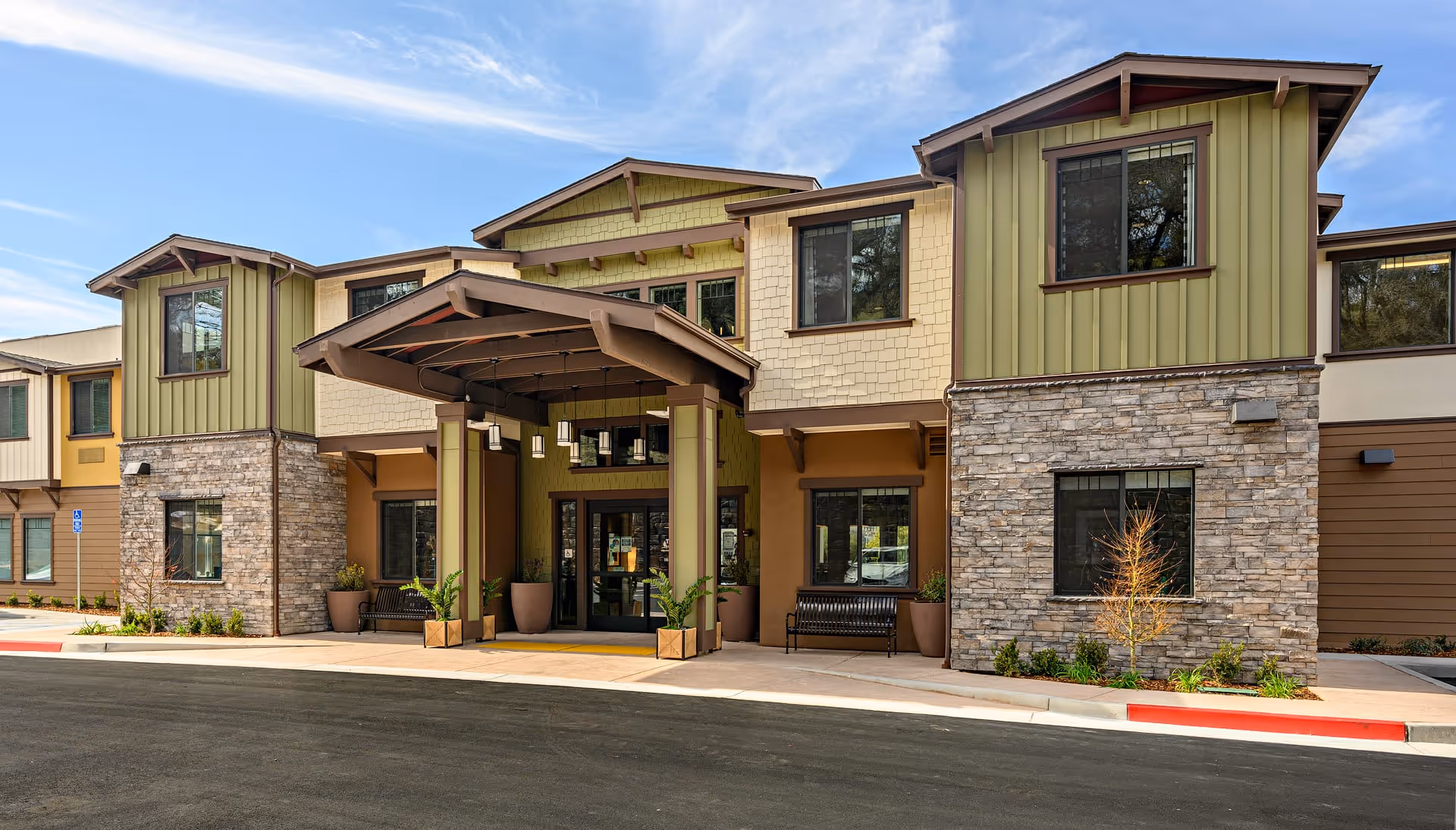 Exterior front entrance of The Oaks at Paso Robles senior living facility featuring a covered drop-off area with hanging lights, stone and green siding, large windows, benches, and landscaped plants under a partly cloudy sky.
