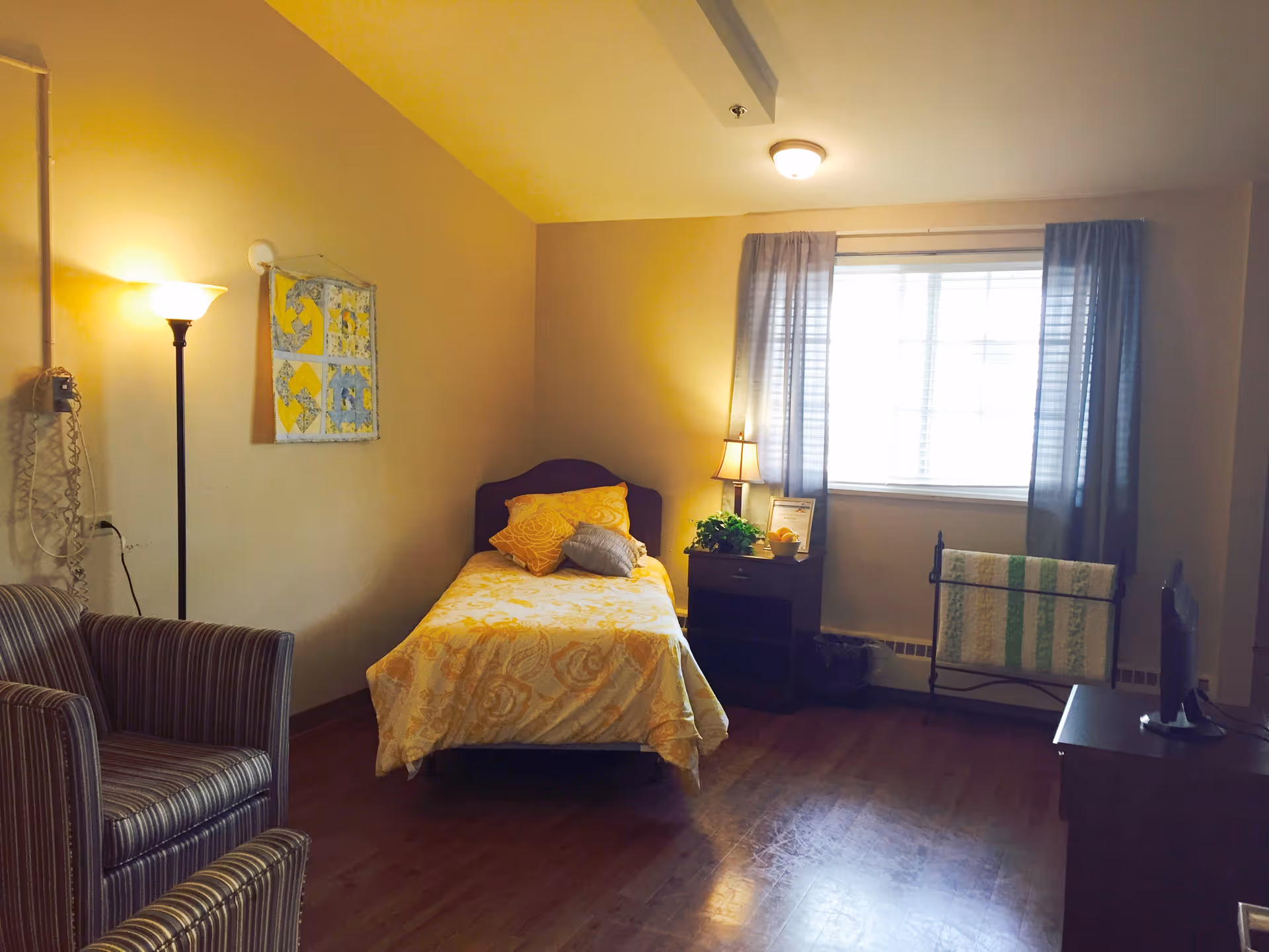 A small furnished bedroom with a single bed, nightstand, armchair, and a window with curtains.
