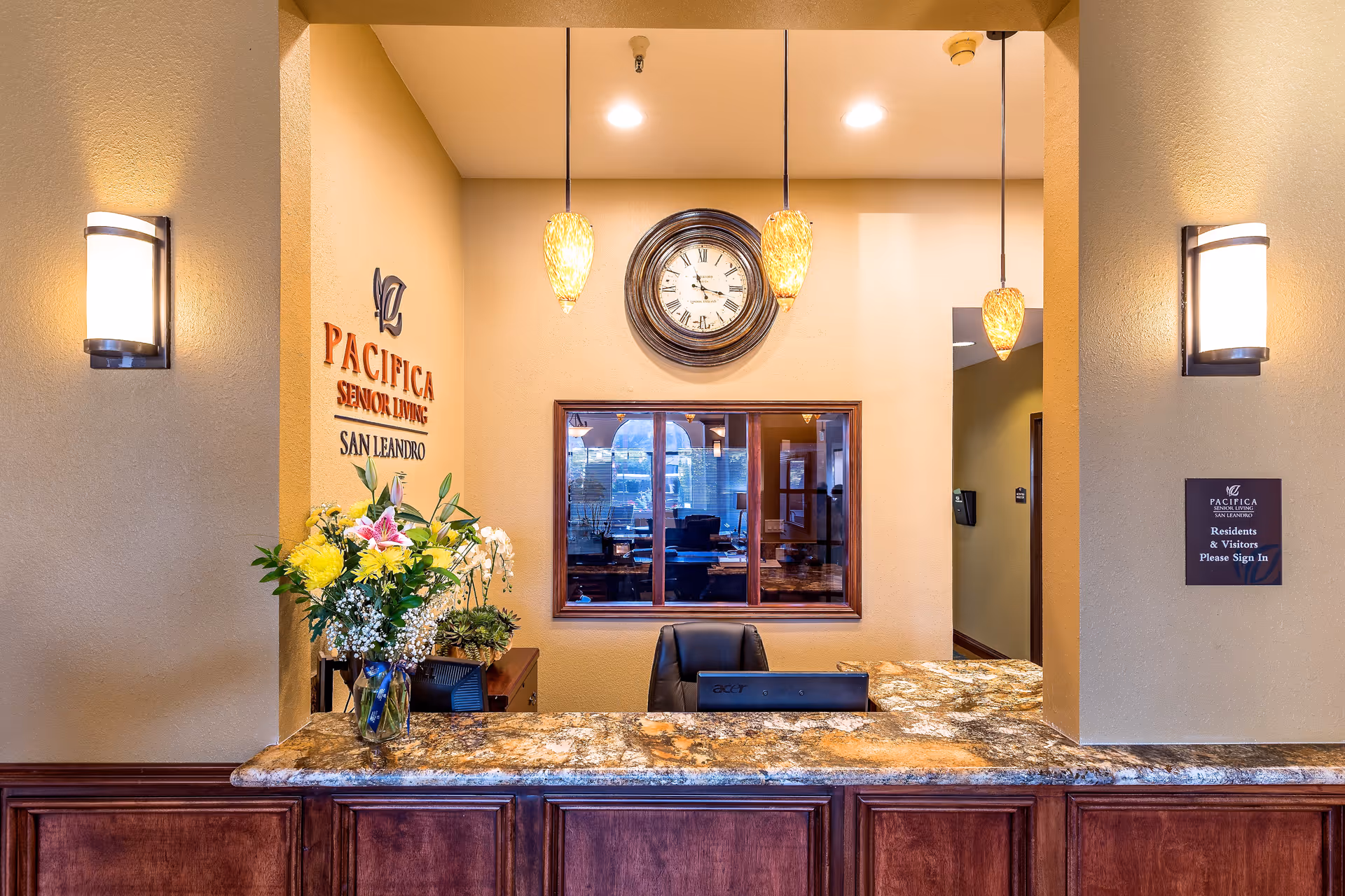 Reception desk area at Pacifica Senior Living San Leandro with a granite countertop, a vase of flowers, a clock on the wall, and pendant lights hanging from the ceiling.